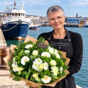 Sur le quai du port de pêche de Boulogne-sur-Mer, la fleuriste Carine Sannier, maître artisan, présente un volumineux bouquet composé de fleurs blanches et de touches verdoyantes enveloppé de papier kraft, avec les chalutiers et la ville en arrière-plan